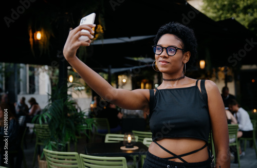 Obraz na płótnie Happy young woman in stylish outfit smiles as she records vlog segment or chats on video call, standing near café tables under cozy lights in relaxed urban setting w salonie