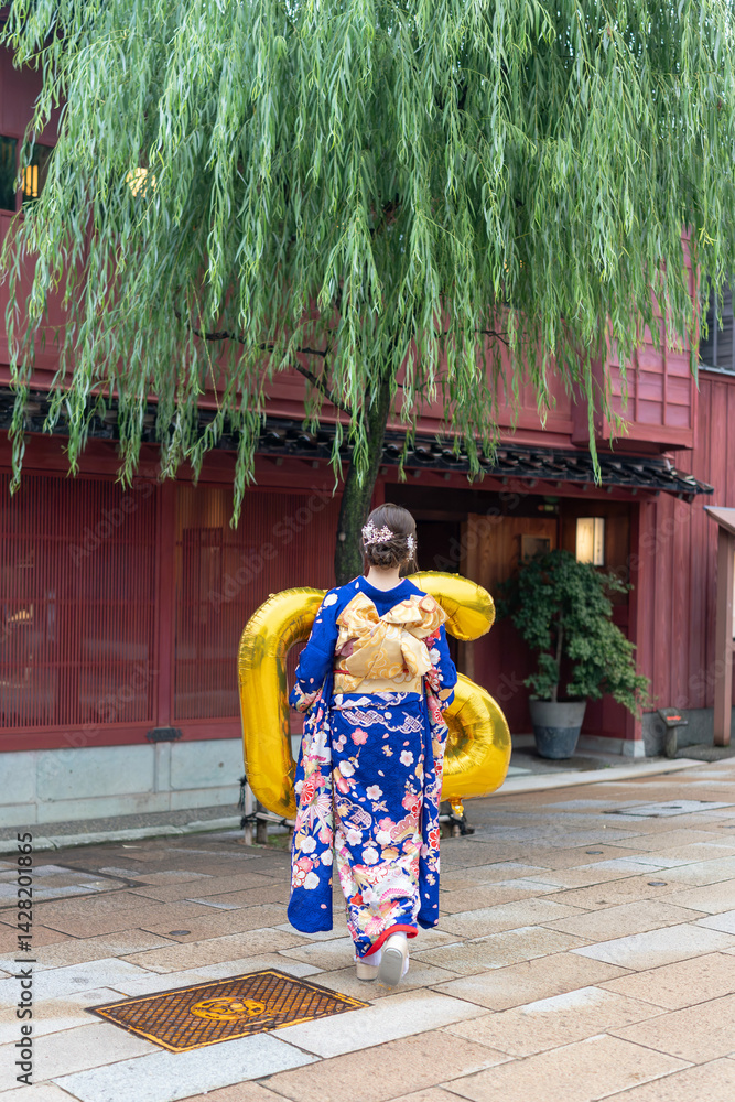 Fototapeta premium On a sunny summer day, a teenage Japanese girl wearing a blue kimono is standing in the traditional townscape of a Japanese city. She is holding large golden balloons shaped like the numbers 2 and 0.