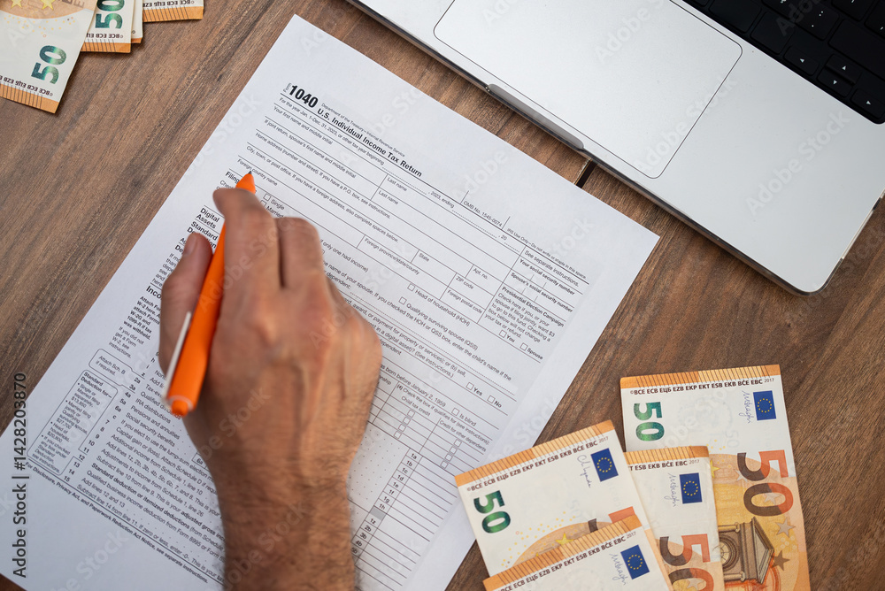 Obraz premium Business owner fills 1040 tax form with pen documenting income and deductions to determine potential refund sitting at table with euro banknotes and laptop