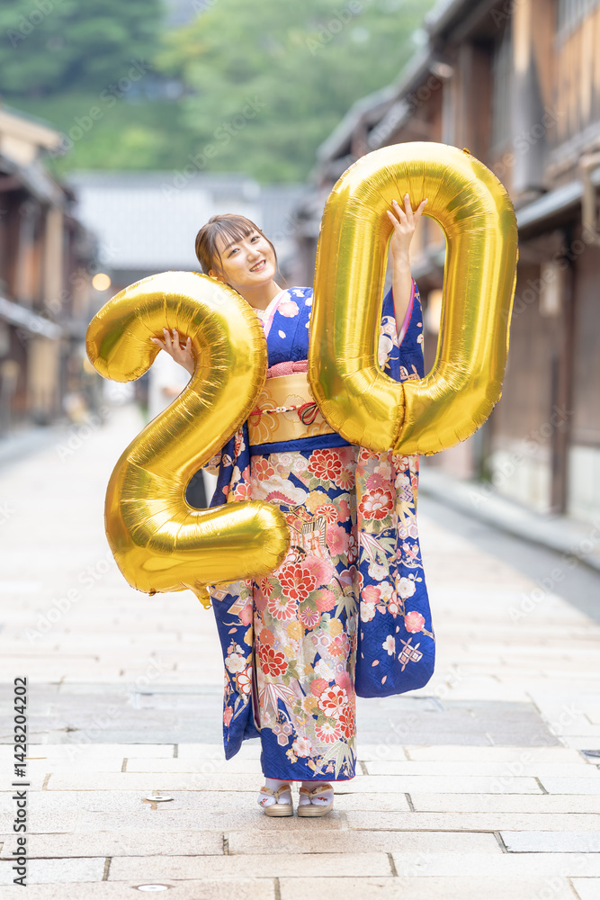 On a summer day, a 20-year-old Japanese girl wearing a blue kimono is in the traditional townscape of a Japanese city. She is holding large golden balloons shaped like the numbers 2 and 0.