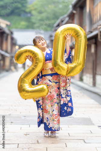 Obraz na płótnie On a summer day, a 20-year-old Japanese girl wearing a blue kimono is in the traditional townscape of a Japanese city. She is holding large golden balloons shaped like the numbers 2 and 0. w salonie