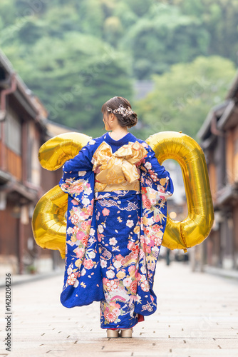 Obraz na płótnie On a summer day, a 20-year-old Japanese girl in a blue kimono stands in a traditional townscape of a Japanese city, holding golden balloons shaped like 2 and 0. Back view. w salonie