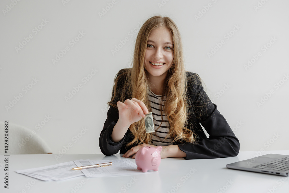 Young woman saving money in a pink piggy bank while smiling at a workstation with documents and a laptop