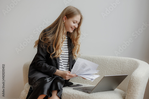 Young woman reviewing documents while using laptop on a cozy chair in a light-filled room