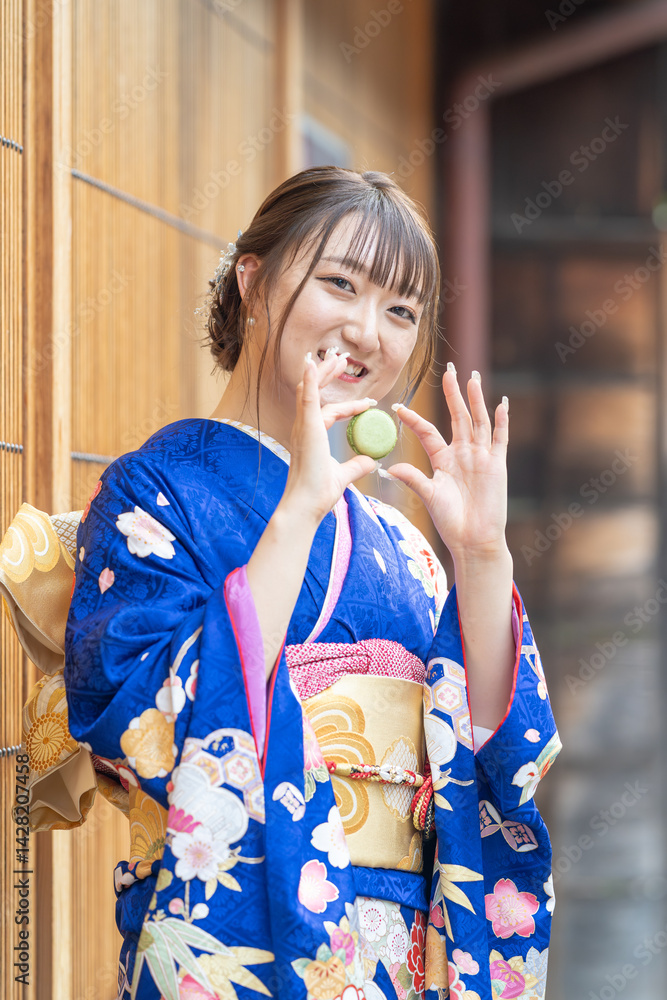 On a summer day, a 20-year-old Japanese girl wearing a blue kimono is standing in the traditional townscape of a Japanese city, holding a green macaron.