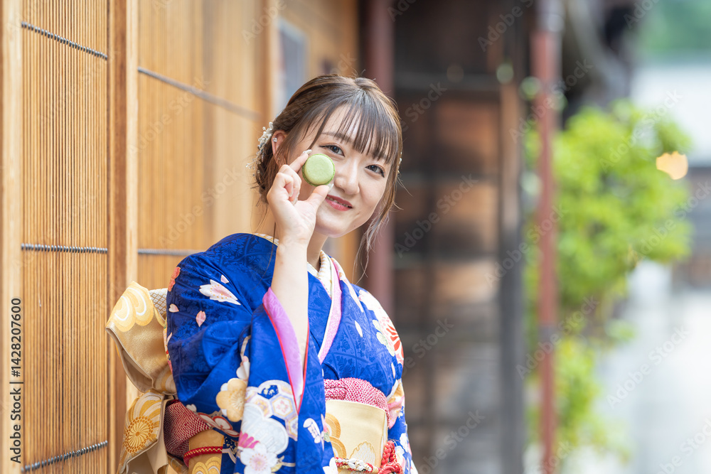 On a summer day, a 20-year-old Japanese girl wearing a blue kimono is standing in the traditional townscape of a Japanese city, holding a green macaron.