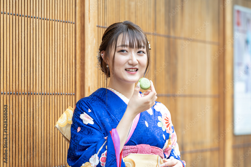 On a summer day, a 20-year-old Japanese girl wearing a blue kimono is standing in the traditional townscape of a Japanese city, holding a green macaron.