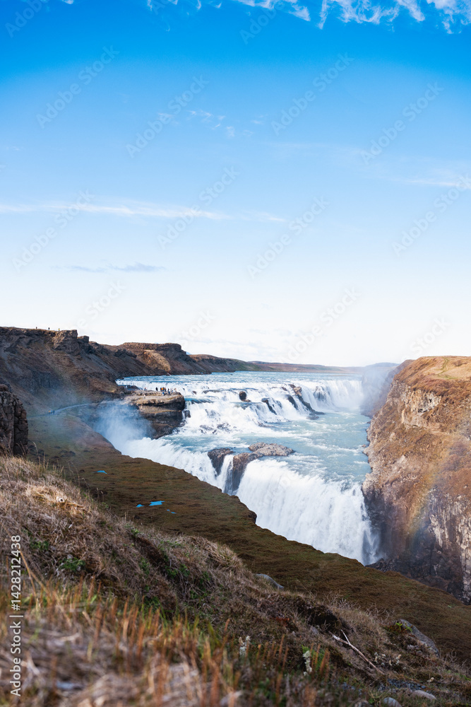 Panoramic view of Gullfoss waterfall cutting through rugged Icelandic cliffs, with turquoise waters and clear skies over the highland plateau.