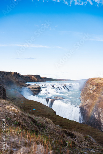 Obraz na płótnie Panoramic view of Gullfoss waterfall cutting through rugged Icelandic cliffs, with turquoise waters and clear skies over the highland plateau. w salonie