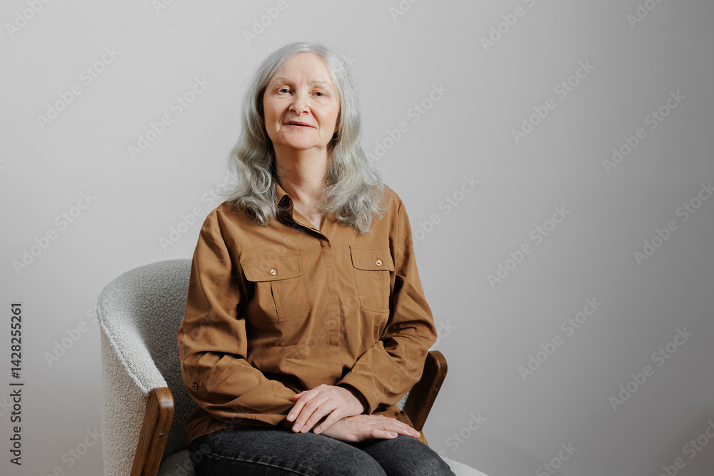 Elderly woman with gray hair sitting comfortably in a casual outfit in a simple indoor setting