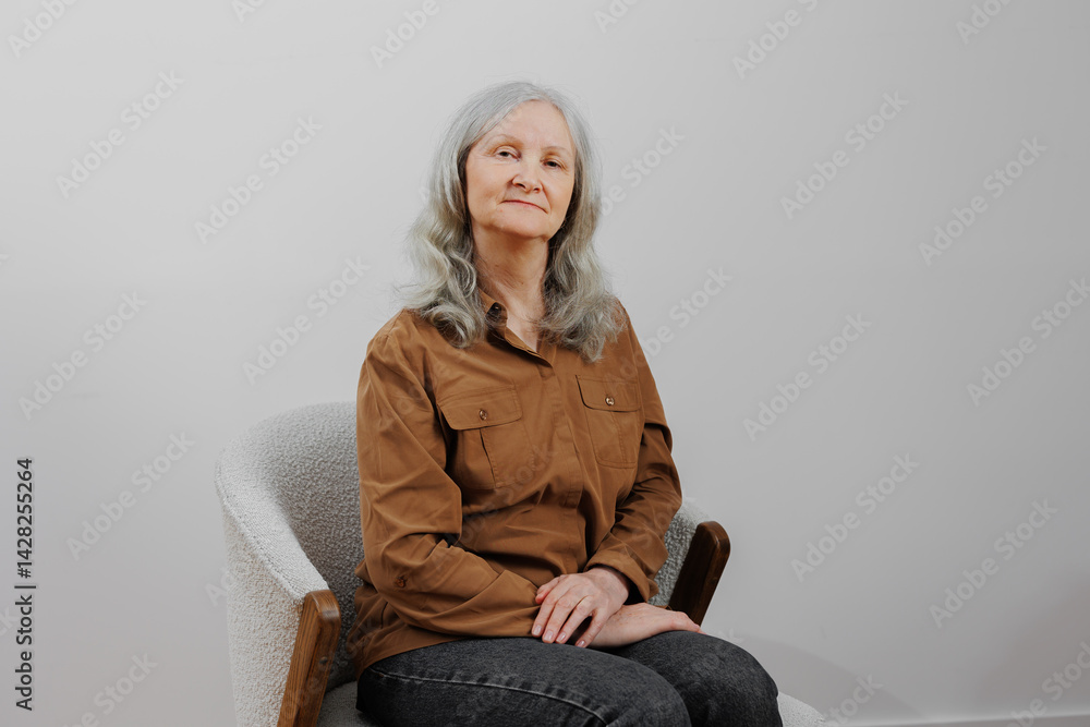 Elderly woman with gray hair sits confidently in a minimalist chair against a neutral background