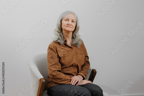 Obraz na płótnie Elderly woman with gray hair sits confidently in a minimalist chair against a neutral background w salonie