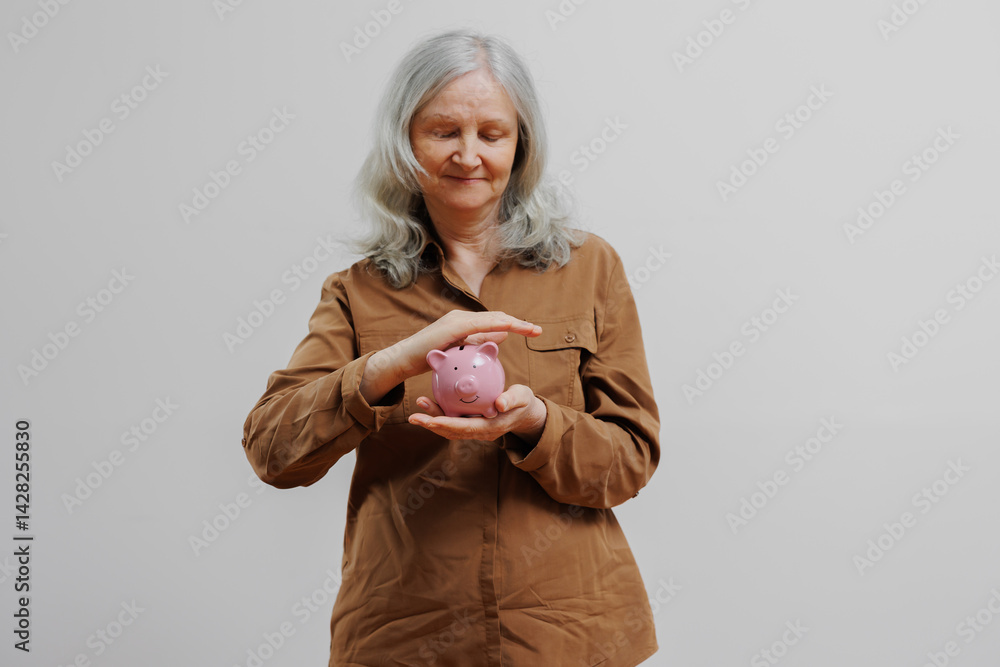 Older woman holding a pink piggy bank and smiling while thinking about saving money in a neutral setting