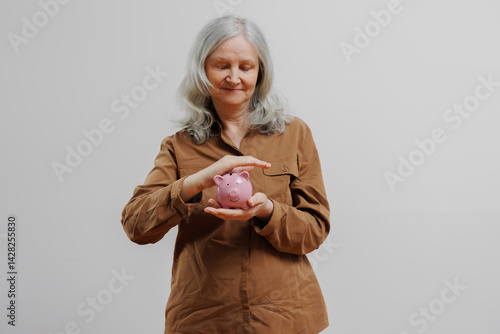 Obraz na płótnie Older woman holding a pink piggy bank and smiling while thinking about saving money in a neutral setting w salonie