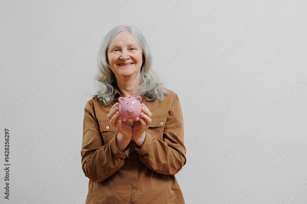 Elderly woman smiles while holding a pink piggy bank indoors during daylight hours in a neutral-colored room