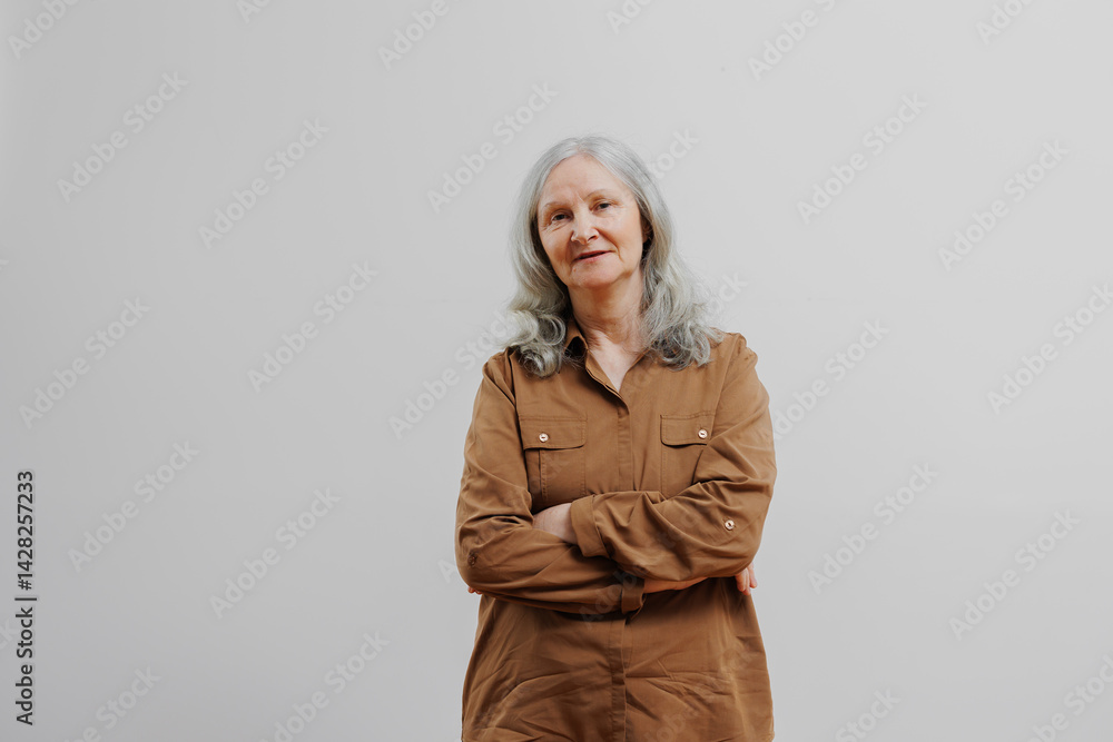 Elderly woman with light gray hair stands confidently with arms crossed in a neutral room during daylight