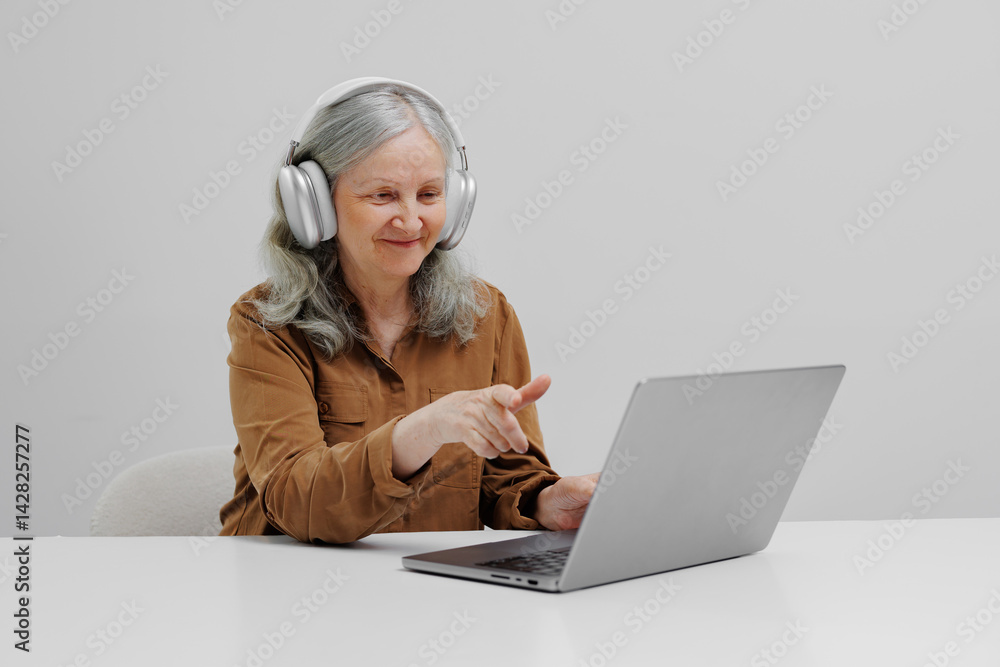 Senior woman wearing headphones engages with laptop in bright, minimalistic space during online activity