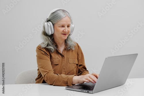 Obraz na płótnie Senior woman using laptop with headphones in a modern workspace during daytime for online activities w salonie