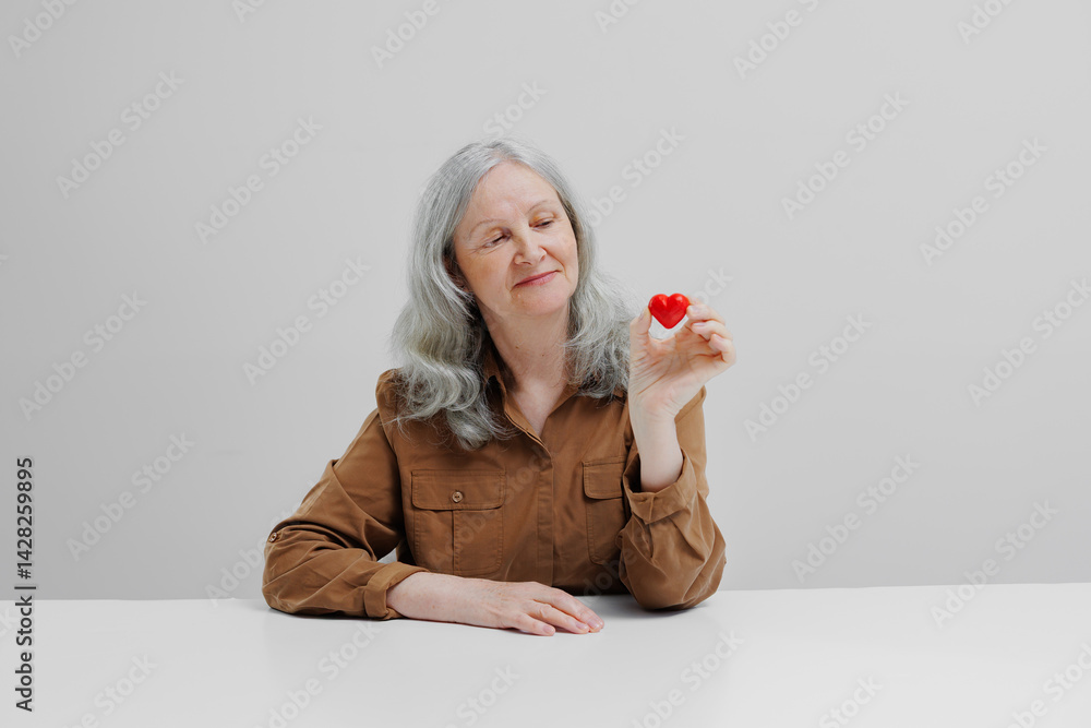 Senior woman smiling while holding a red heart figurine in a minimalist setting