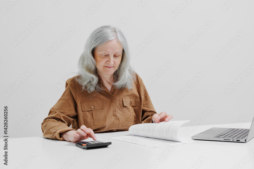 Older woman studying financial documents while using a calculator at a white desk near a laptop