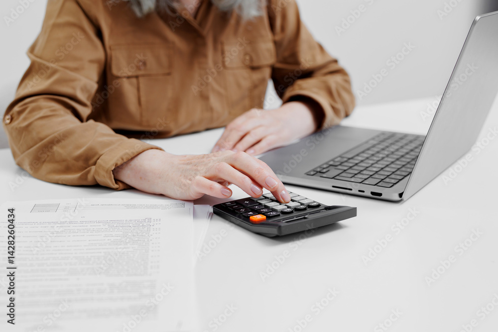 Person working on laptop while calculating numbers using a calculator in a minimalistic office environment