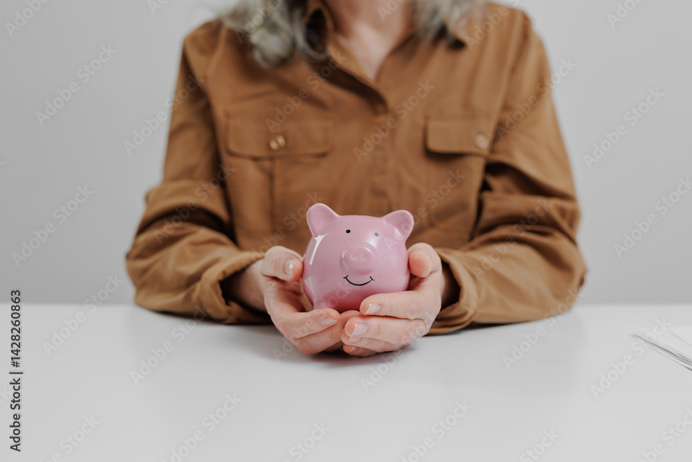 Person holding a pink piggy bank while seated at a table in a neutral workspace