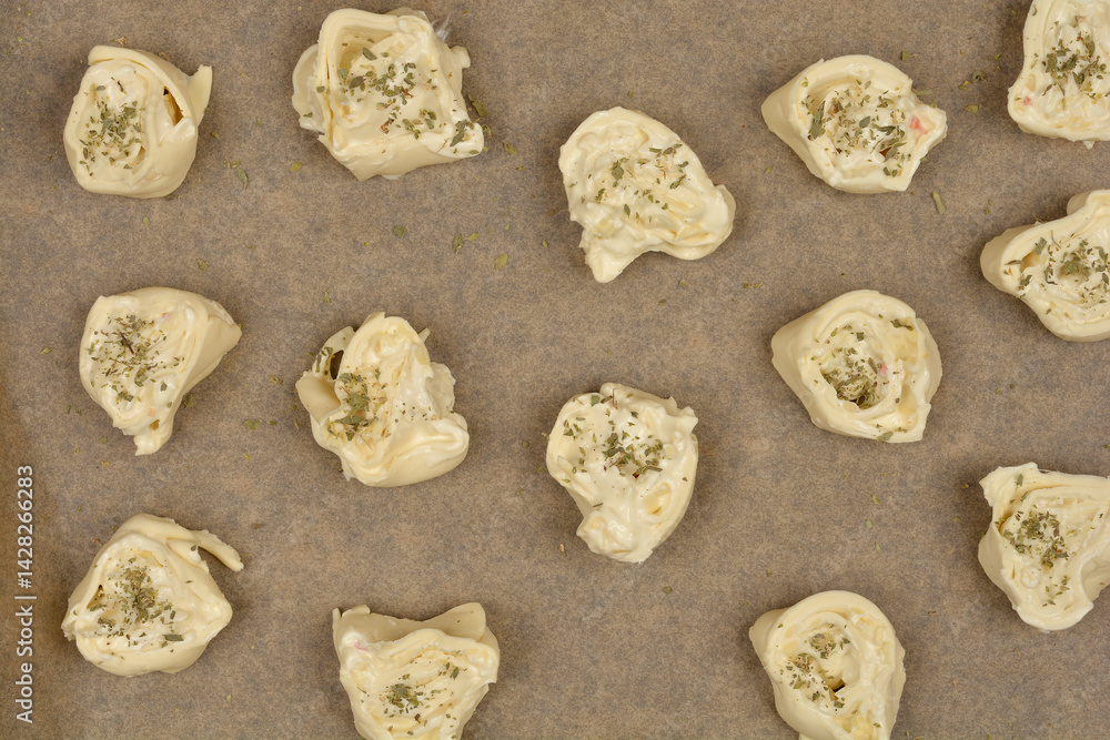 An overhead shot shows raw cheese and oregano puff pastry rolls arranged on parchment paper on a baking sheet, ready to be baked