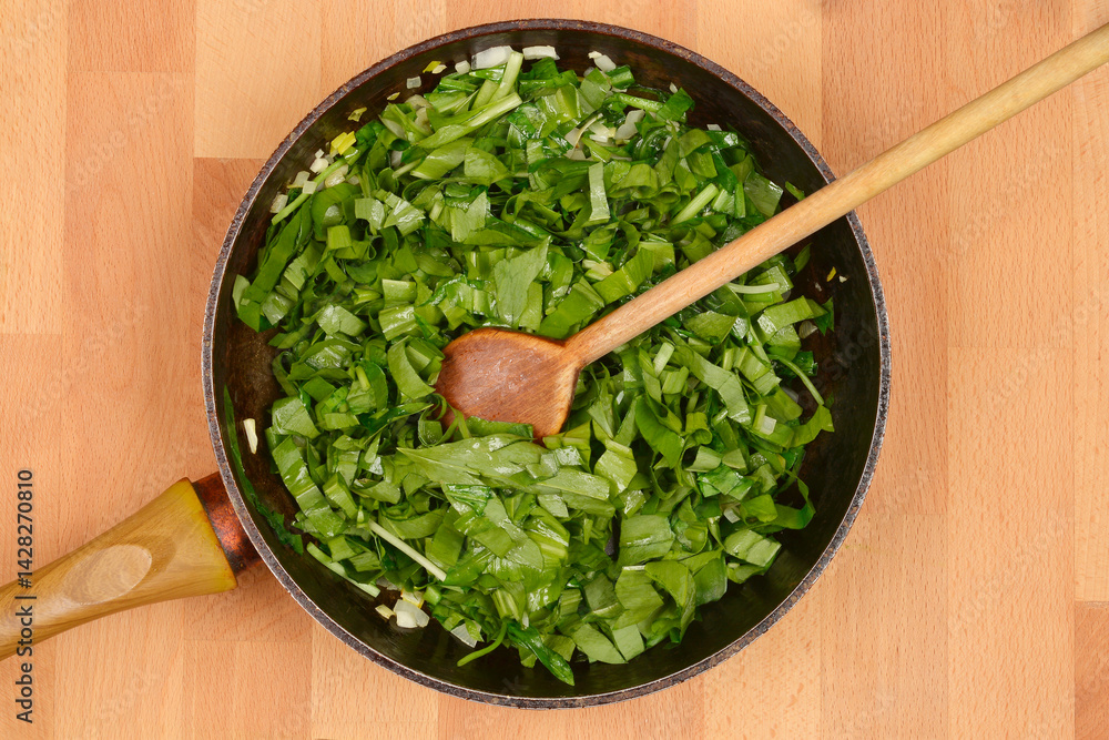 Chopped wild garlic or bears garlic (Allium ursinum) leaves and white onions are being saued in a black frying pan with a wooden spoon resting inside