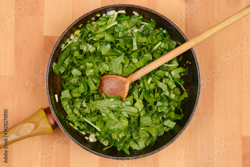 Chopped wild garlic or bears garlic (Allium ursinum) leaves and white onions are being saued in a black frying pan with a wooden spoon resting inside