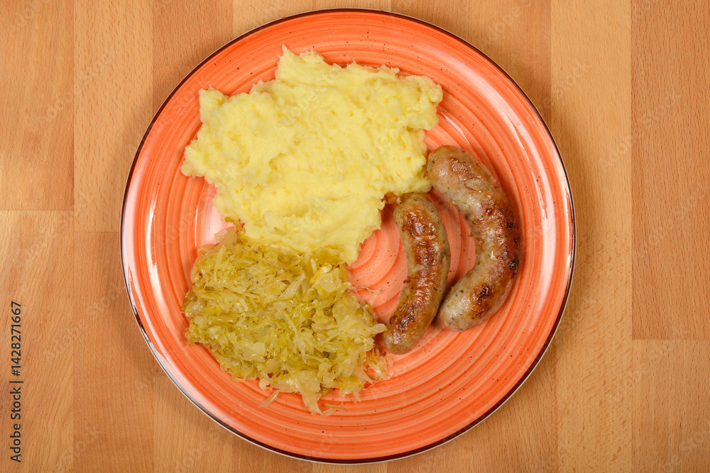 A plate features two grilled homemade sausages served with mashed potatoes and sauerkraut on a light wooden surface