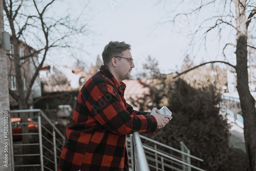 Obraz na płótnie A young blond man wearing glasses, looking at his phone while walking down the street. A modern scene showcasing urban life, technology, and casual street style w salonie