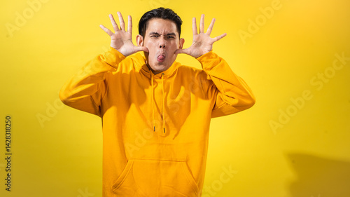 Tableau sur toile Asian man in yellow hoodie making a silly face by pulling cheeks and sticking out tongue, with both hands raised against a bright yellow background