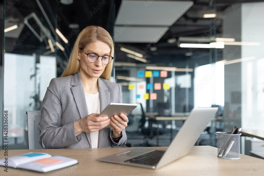 A businesswoman with glasses uses a tablet while working at a desk in a modern office. A laptop and notebook are also present. A businesswoman with glasses uses a tablet while working at a desk in a modern office. A laptop and notebook are also present.