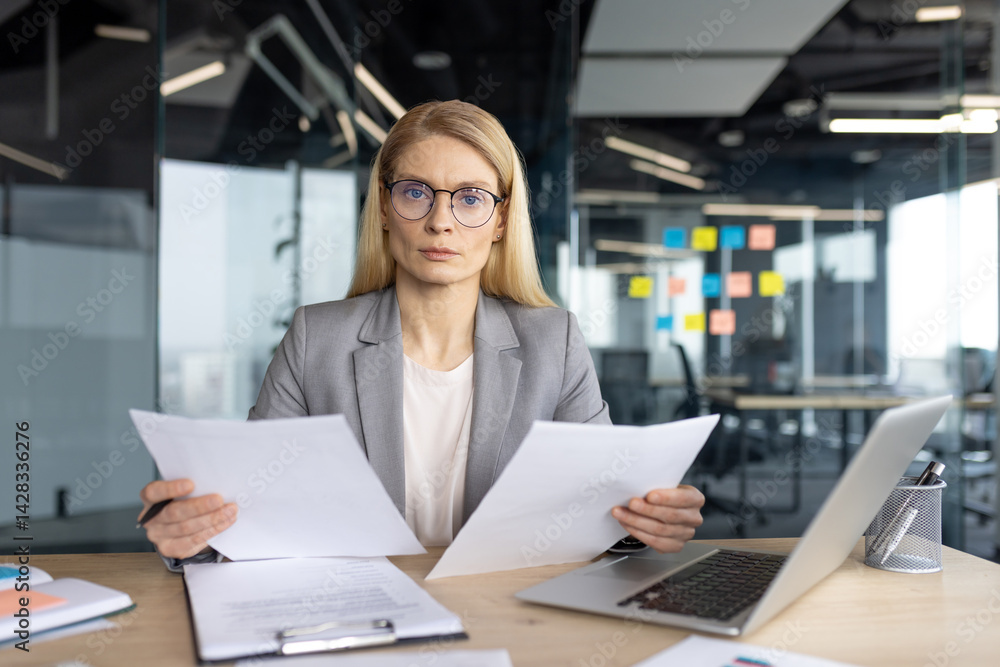 A businesswoman in a professional setting sits at her desk, reviewing documents with a serious expression. She is wearing glasses.