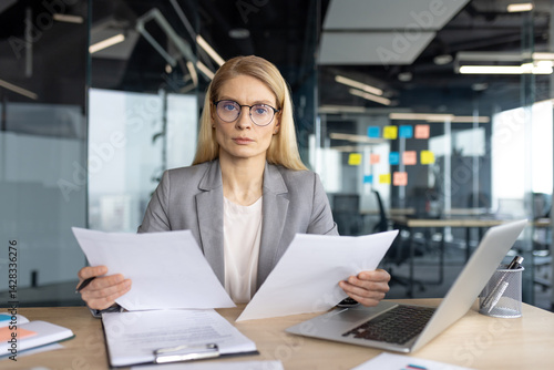 Obraz na płótnie A businesswoman in a professional setting sits at her desk, reviewing documents with a serious expression. She is wearing glasses. w salonie