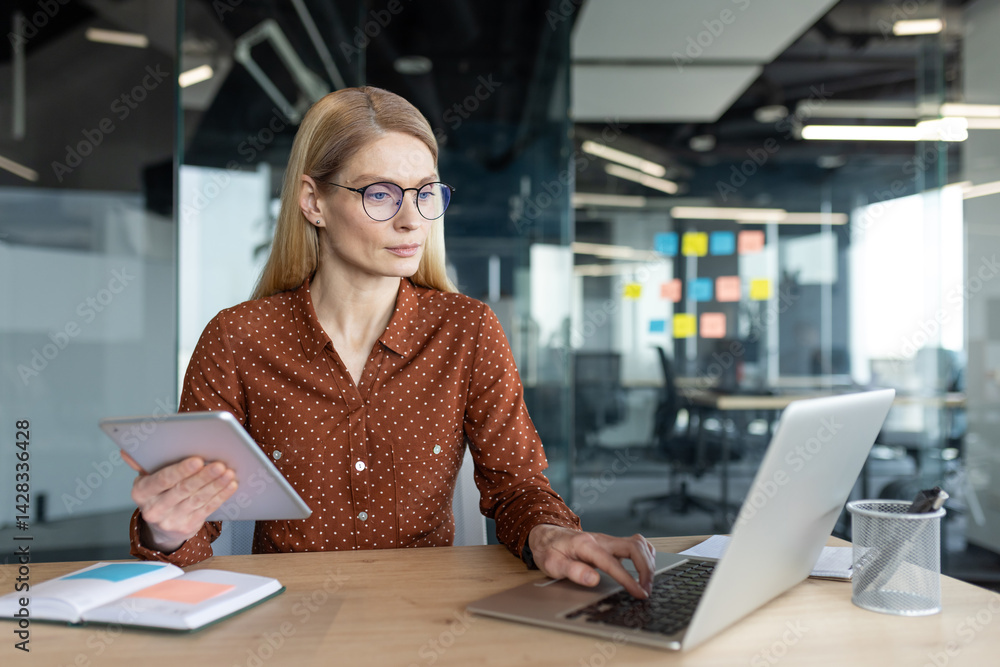 A businesswoman works on her laptop in a modern office, holding a tablet and looking thoughtfully. She wears glasses.