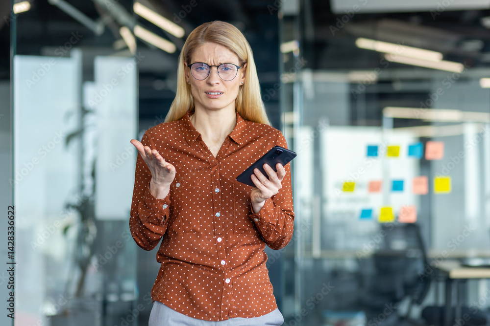 A frustrated woman in glasses holds a phone, gesturing with confusion in an office setting with glass walls and colorful sticky notes.