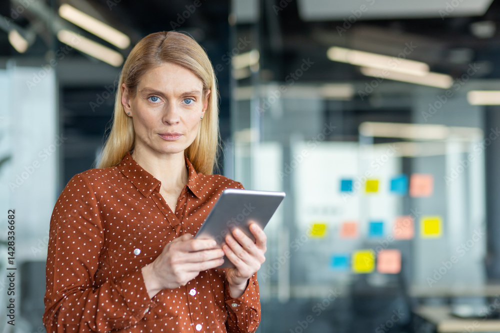 A poised businesswoman in a modern office setting, holding a digital tablet, looking directly at the camera with a composed expression.
