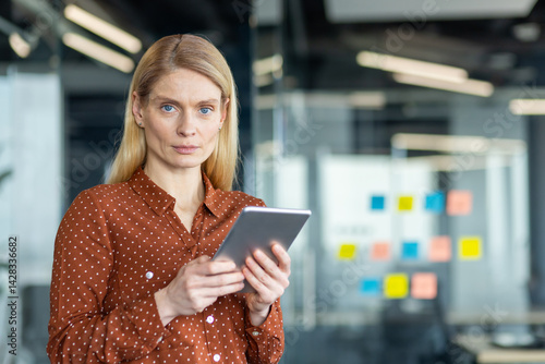 Obraz na płótnie A poised businesswoman in a modern office setting, holding a digital tablet, looking directly at the camera with a composed expression. w salonie