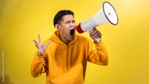 Young Asian man in yellow hoodie yelling into a red and white megaphone with eyes closed, showing strong emotion against bright yellow background. Concept of protest, urgency, or bold announcement