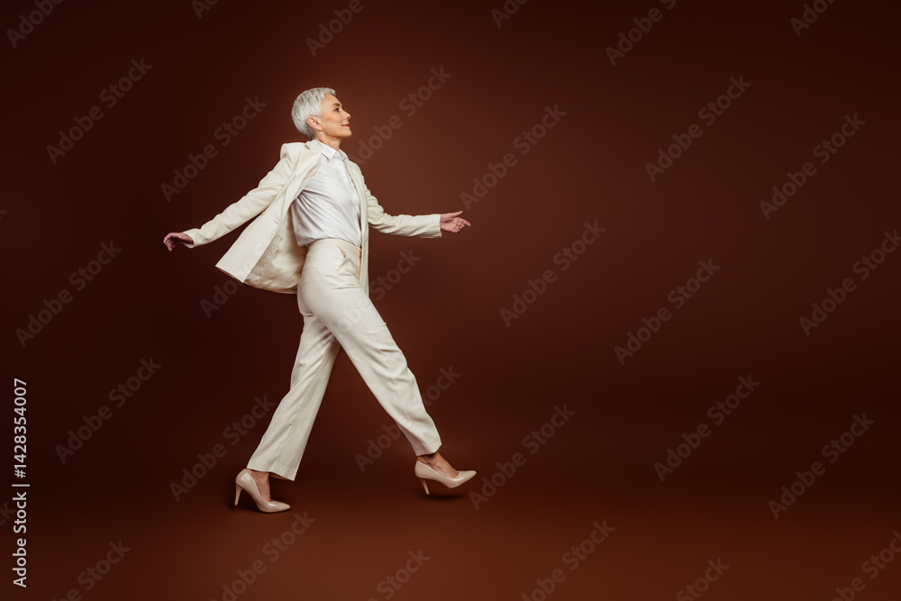 Confident mature professional woman posing in a white formal suit against a brown background, showcasing elegance, success Confident mature professional woman posing in a white formal suit against a brown background, showcasing elegance, success