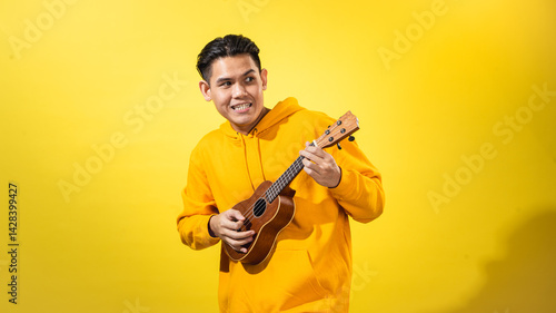 Asian man in yellow hoodie enthusiastically playing a small ukulele against a vivid yellow background. Expressive musical portrait with happy mood and creative, artistic personality