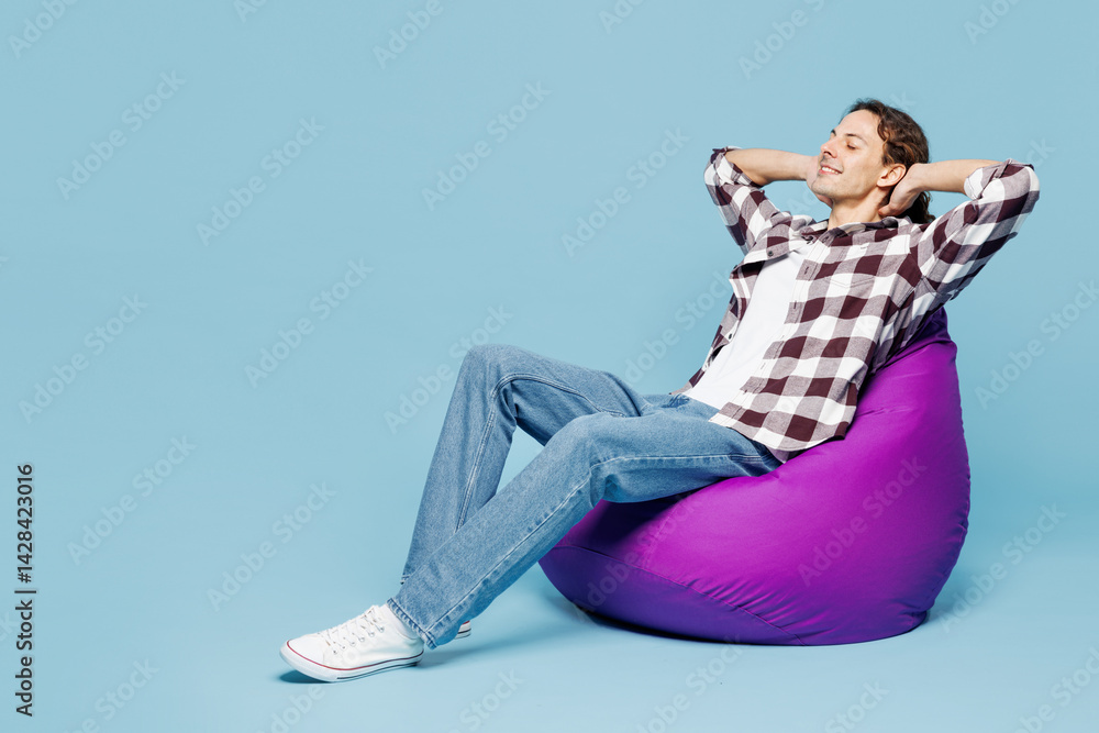 Obraz premium Full body young calm man he wear purple checkered shirt casual clothes sit in bag chair hold hands behind neck isolated on plain pastel light blue cyan background studio portrait. Lifestyle concept.