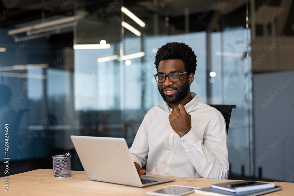 A smiling Black businessman in glasses sits at a desk with a laptop, ready for work. Modern office with glass walls is visible in the background.