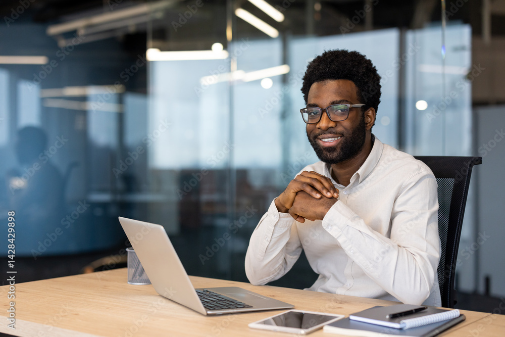 A smiling black businessman sits at his office desk with a laptop, notebook, and tablet, looking content.