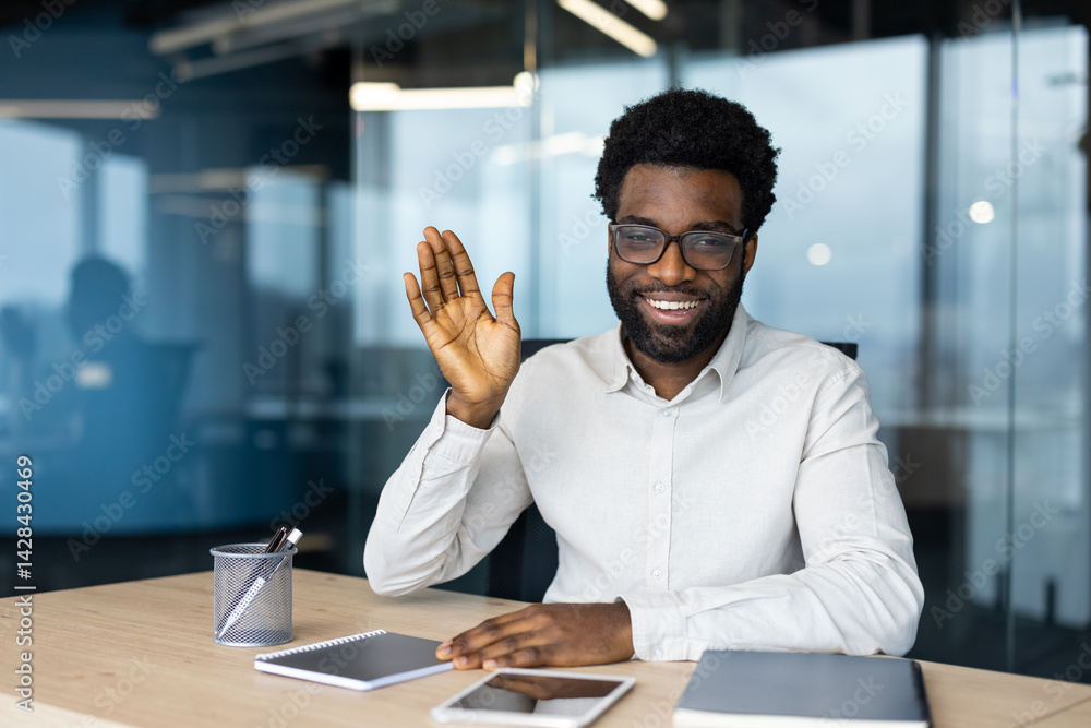 Smiling, well-dressed businessman with glasses waving his hand in an office setting, greeting during a virtual meeting or video call.