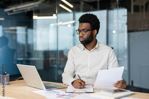 An african american businessman is at his desk, working on some documents in an office environment. A laptop and tablet are nearby.