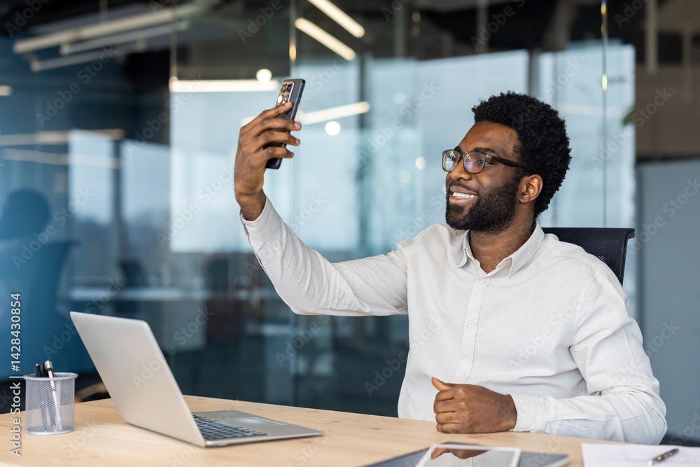 A smiling businessman takes a selfie in his modern office. He is wearing glasses and a white shirt, enjoying the moment.