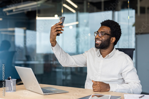 A smiling businessman takes a selfie in his modern office. He is wearing glasses and a white shirt, enjoying the moment.