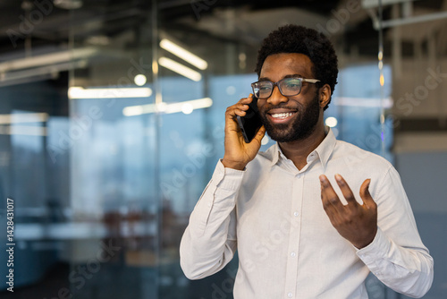 Obraz na płótnie A cheerful businessman with glasses chats on his phone in an office, likely discussing business matters. He seems happy during the call. w salonie